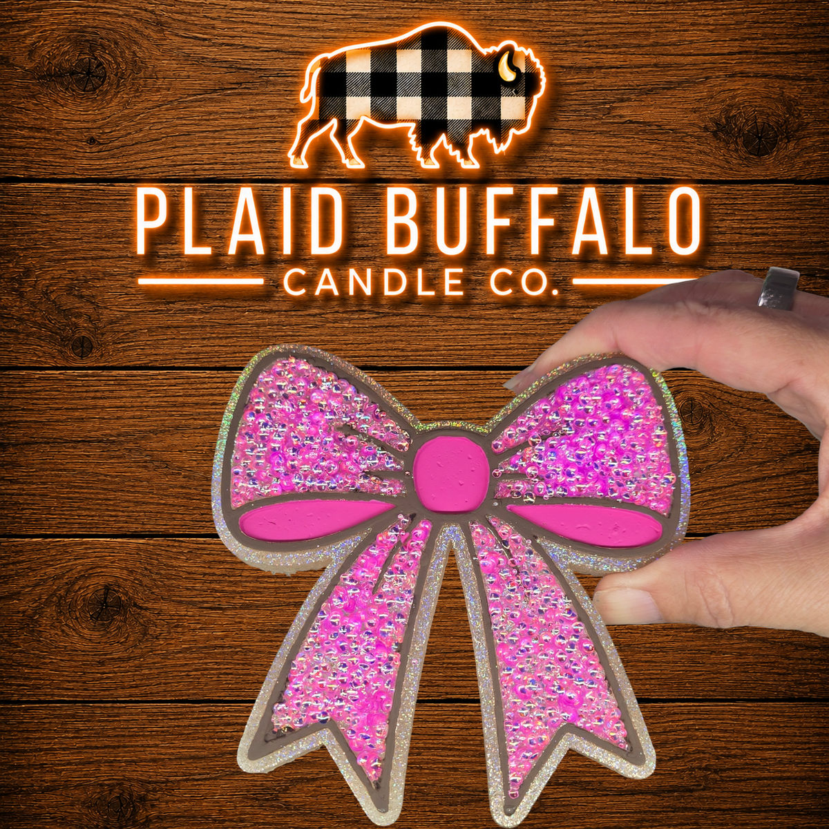 Large bow-shaped car freshie featuring hot pink accents, iridescent pink rhinestones, and a sparkling silver glitter outline, displayed on a rustic wood background with the Plaid Buffalo Candle Co. logo.