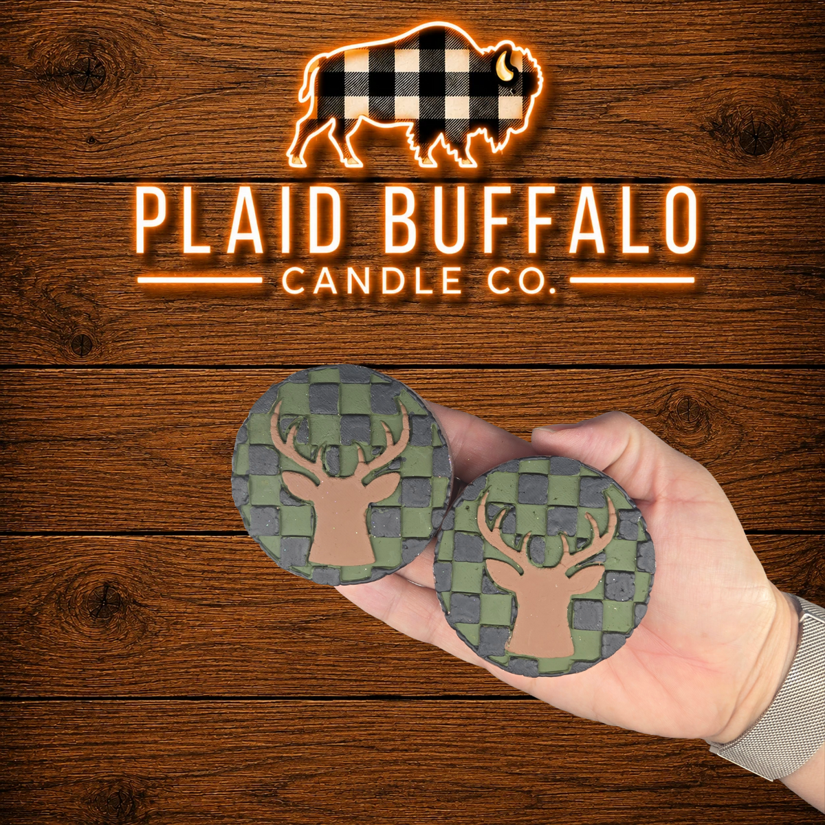 Pair of handmade deer head vent clips with a green and charcoal buffalo plaid background, displayed in hand on a rustic wood backdrop.
