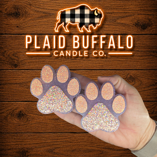 Pair of handmade glittery paw print vent clips in lavender with sparkling pink and iridescent accents, displayed in hand on a rustic wood background.
