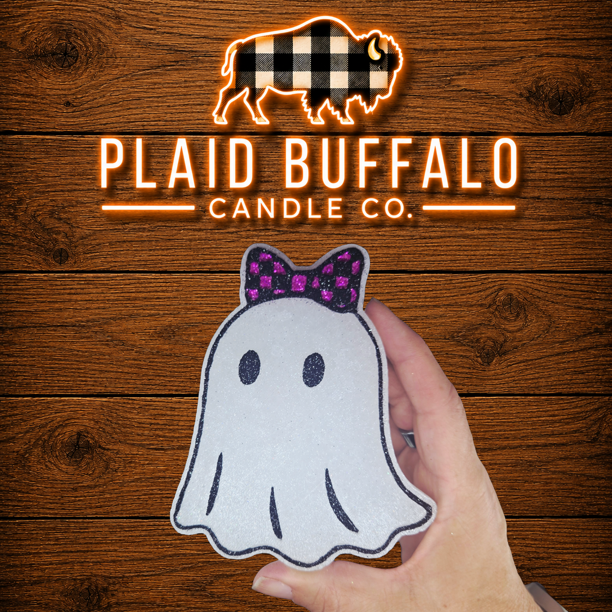 A hand holds a white scented ghost-shaped car freshener with a purple bow against a rustic wood plank background, with the Plaid Buffalo Candle Co. logo displayed above.
