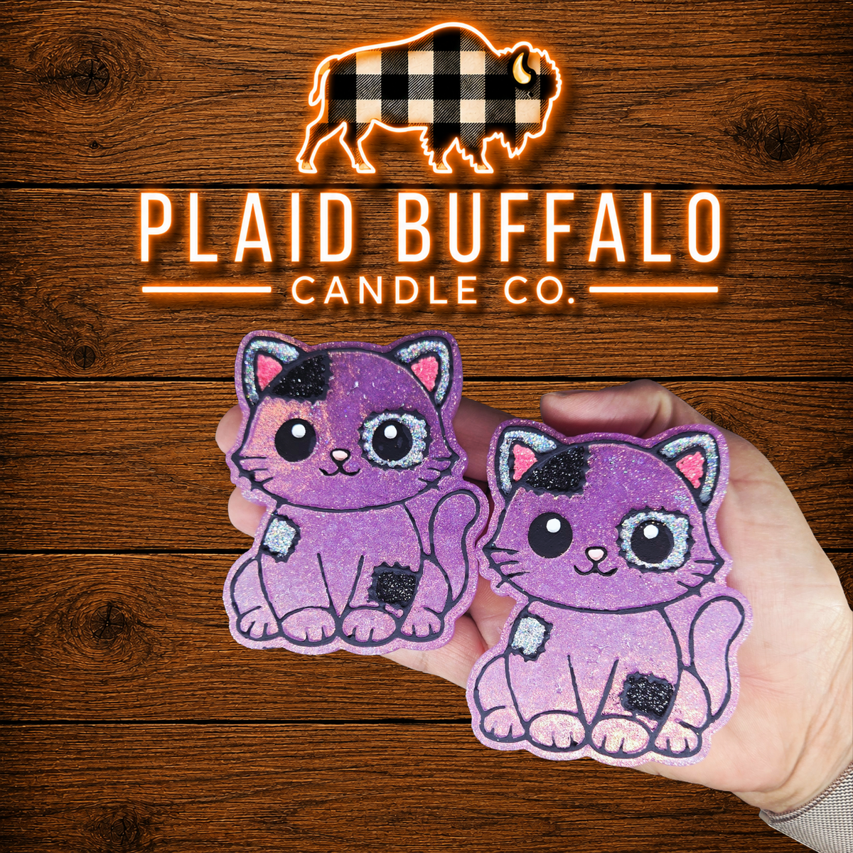 Hand holding two purple glitter patchwork cat-shaped scented car freshies with black, silver, and pink details against a rustic wood background with the Plaid Buffalo Candle Co. logo.