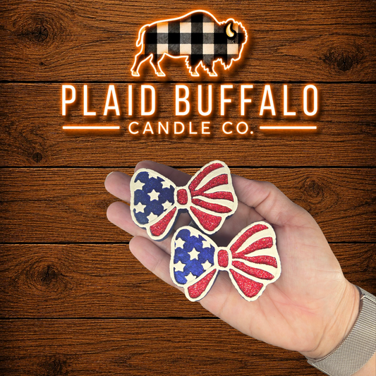 Pair of handmade patriotic bow vent clips featuring red glitter stripes, blue stars, and white accents, displayed in hand on a rustic wood background