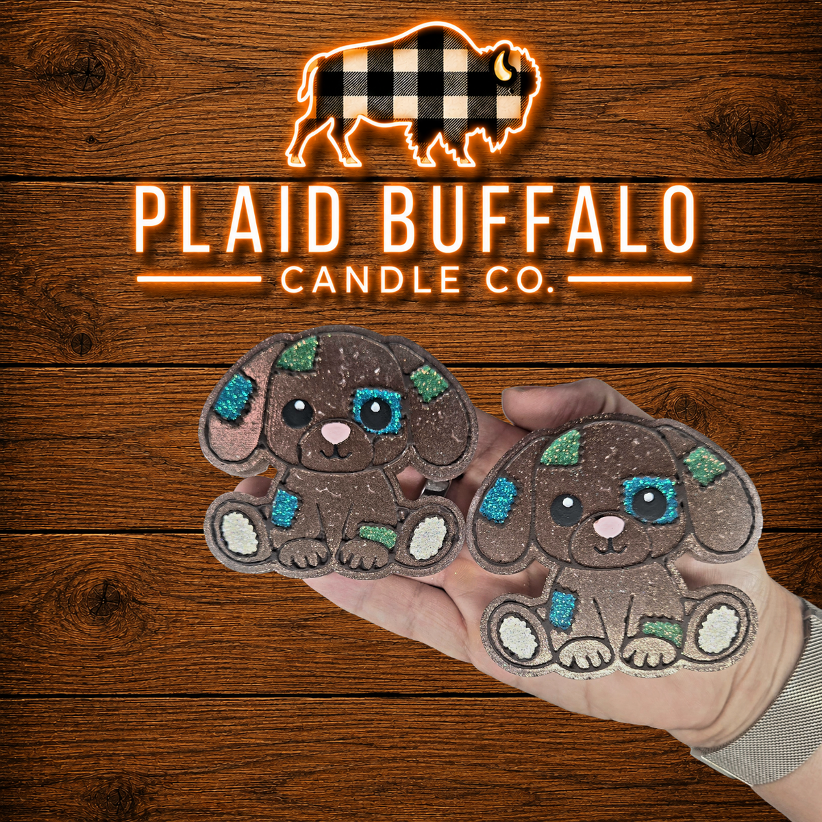A hand holds two small dog-shaped car fresheners in front of a wooden plank background with the brand logo 'PLAID BUFFALO CANDLE CO.' above.