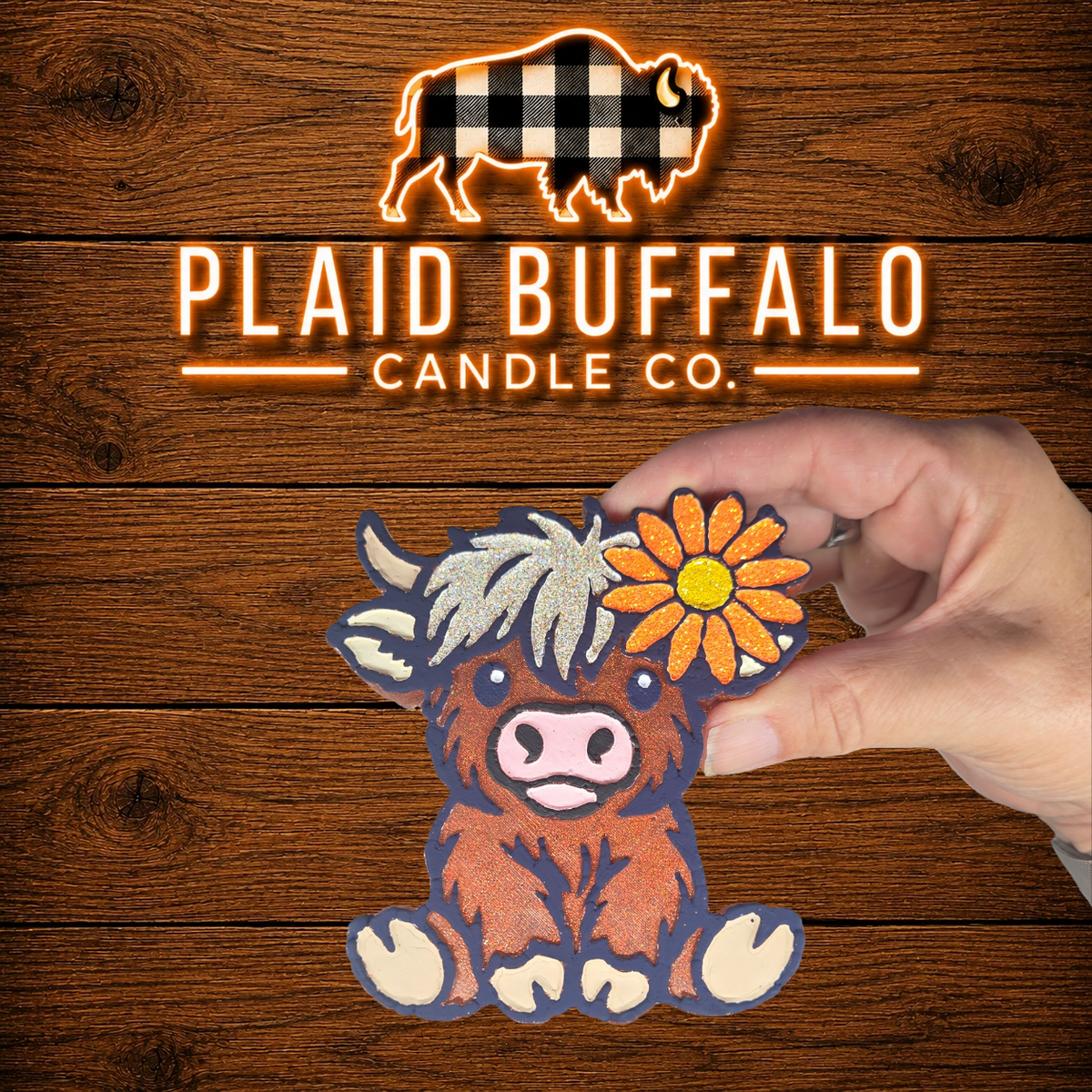 Handmade Highland cow car freshener with a glittery brown coat, silver bangs, and a bright orange sunflower on a rustic wood background.