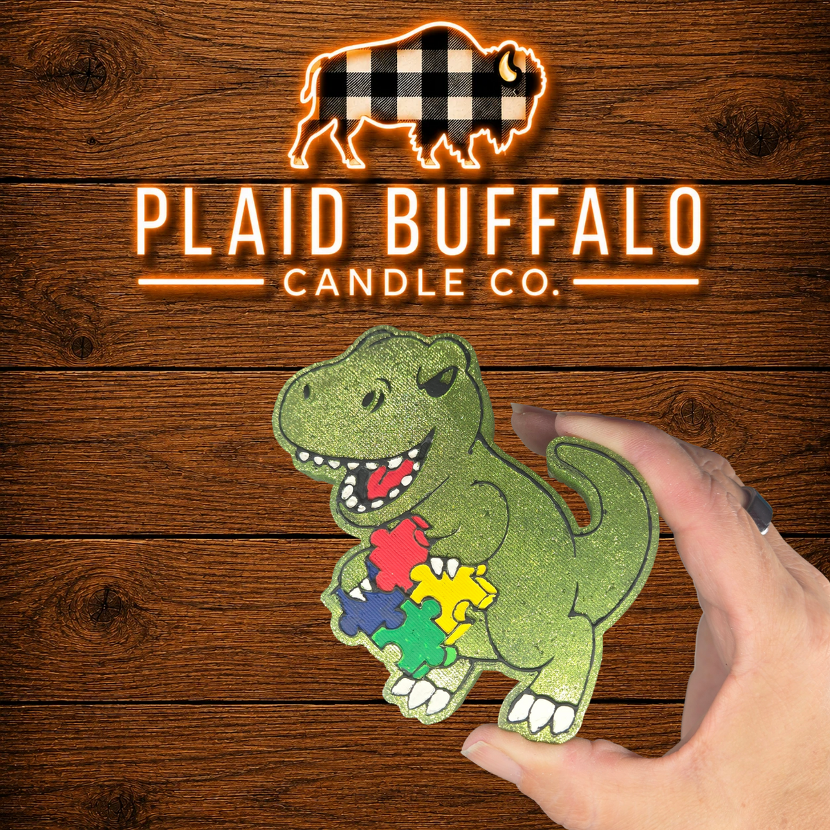 Glittery green T-Rex car freshener holding colorful autism awareness puzzle pieces, handmade and displayed on a rustic wood background.