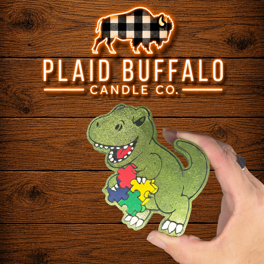 Glittery green T-Rex car freshener holding colorful autism awareness puzzle pieces, handmade and displayed on a rustic wood background.