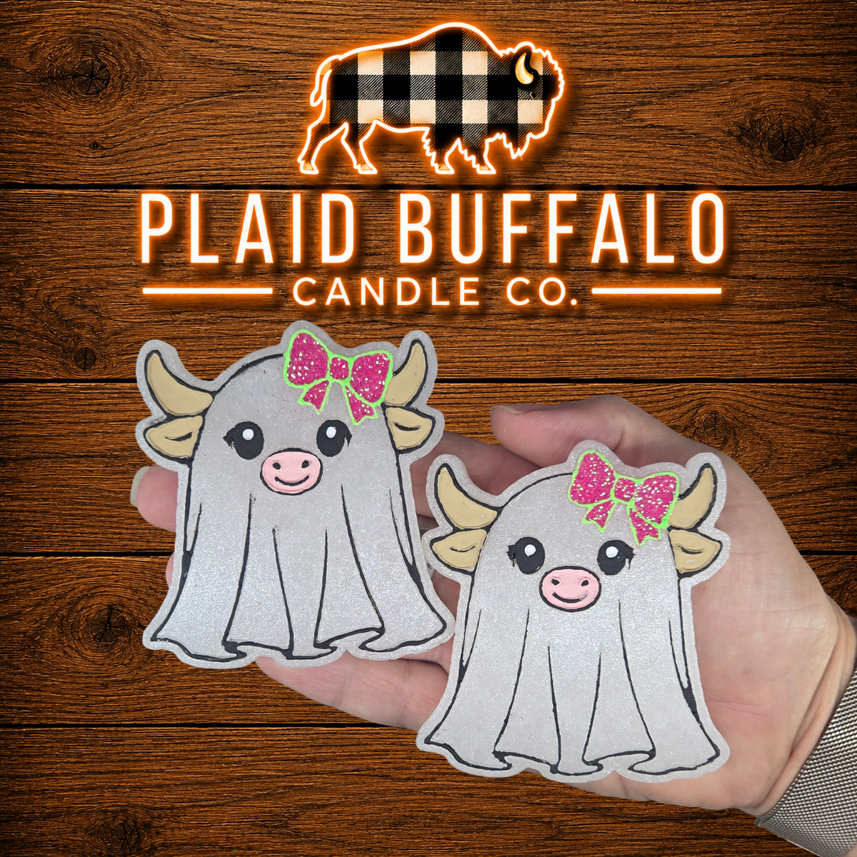Set of two Highland cow ghost-shaped car freshies with soft white ghost bodies, cream-colored horns, pink noses, and bright pink glitter bows, displayed on a rustic wood background with the Plaid Buffalo Candle Co. logo.