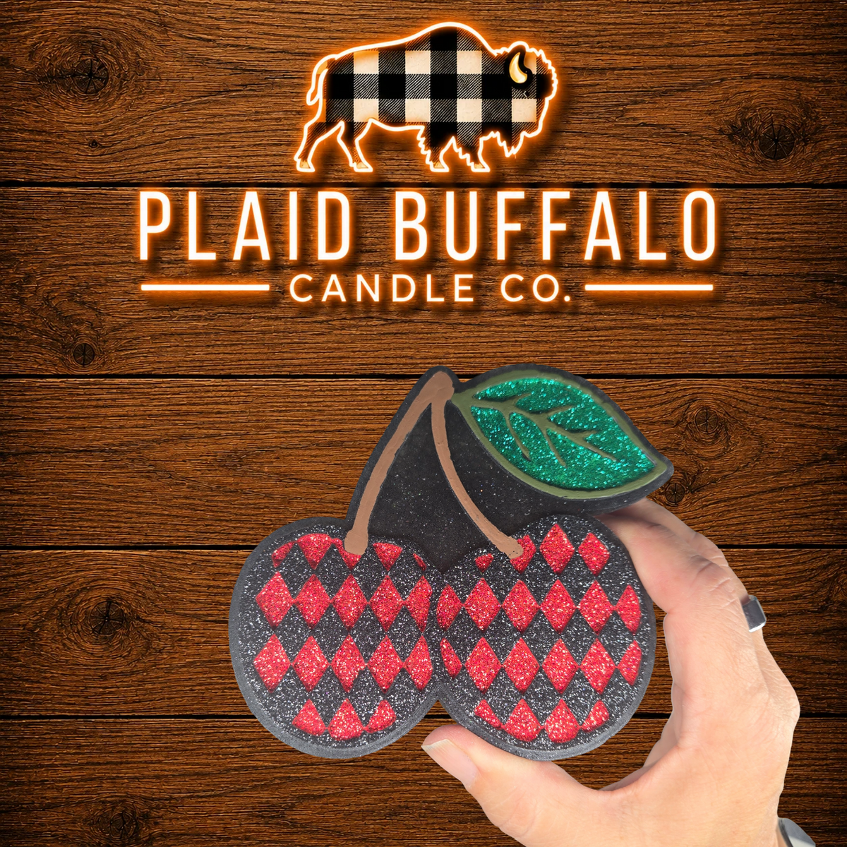 Cherry-shaped car freshener with a red glitter diamond pattern, green glitter leaf, black glitter outline, and rustic wood background with Plaid Buffalo Candle Co. branding.
