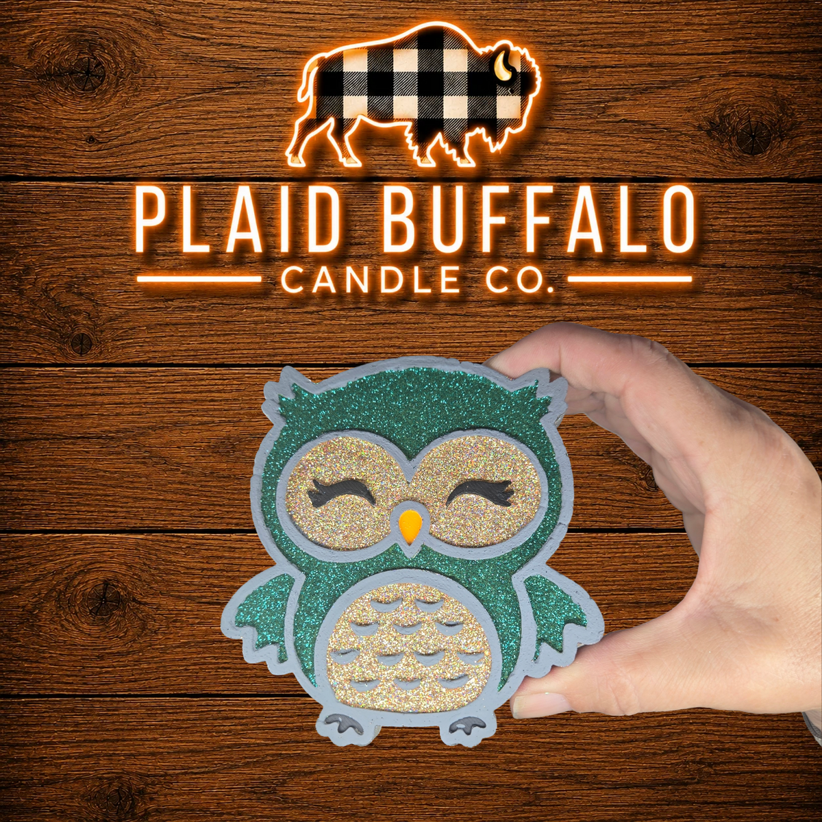 Handmade glittery owl car freshener featuring teal and silver accents with closed eyes and an orange beak, displayed on a rustic wood background.