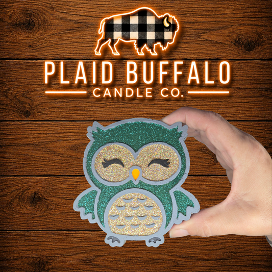 Handmade glittery owl car freshener featuring teal and silver accents with closed eyes and an orange beak, displayed on a rustic wood background.
