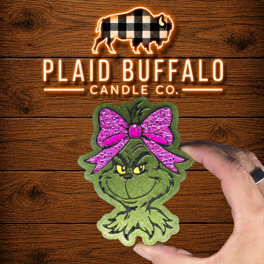 Hand-painted Grinch-inspired car freshie with a shimmering green face, bright yellow eyes, and a large hot pink rhinestone bow, displayed on a rustic wood background with the Plaid Buffalo Candle Co. logo.