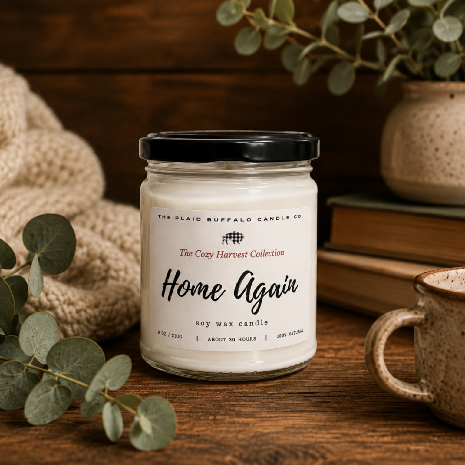 Home Again soy candle by Plaid Buffalo Candle Co. displayed with a cozy knit blanket, eucalyptus stems, stacked books, and ceramic mugs.
