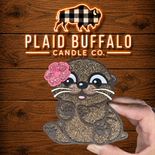 Hand-painted glitter otter car freshie with a pink hibiscus flower, displayed on a rustic wood background with the Plaid Buffalo Candle Co. logo.