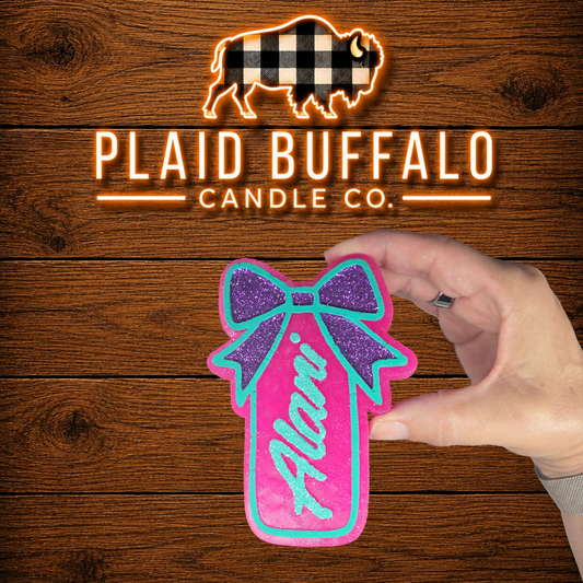 Personalized bright pink car freshener with the name "Alani" in teal glitter lettering and a purple glitter bow, displayed on a rustic wood background.