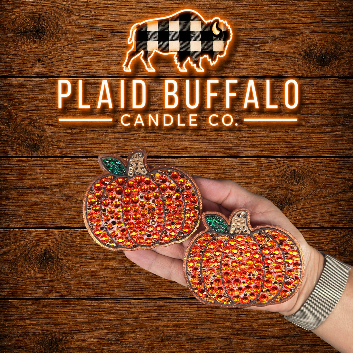 Set of two pumpkin-shaped vent clips featuring sparkling orange rhinestones, bronze glitter accents, green glitter leaves, and textured stems, displayed on a rustic wood background with the Plaid Buffalo Candle Co. logo.