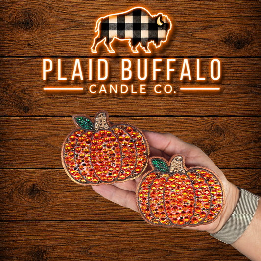 Set of two pumpkin-shaped vent clips featuring sparkling orange rhinestones, bronze glitter accents, green glitter leaves, and textured stems, displayed on a rustic wood background with the Plaid Buffalo Candle Co. logo.