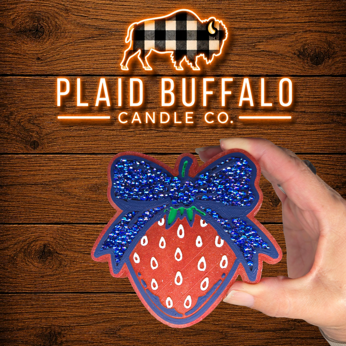Hand-painted strawberry-shaped car freshie featuring a bright red strawberry with white seed details and a large navy blue rhinestone bow, displayed on a rustic wood background with the Plaid Buffalo Candle Co. logo.