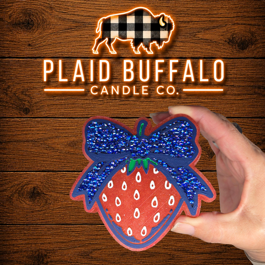 Hand-painted strawberry-shaped car freshie featuring a bright red strawberry with white seed details and a large navy blue rhinestone bow, displayed on a rustic wood background with the Plaid Buffalo Candle Co. logo.