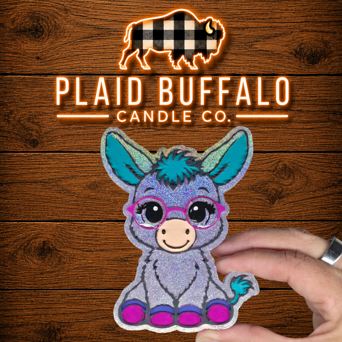 Hand-painted glitter donkey car freshie with teal ears and mane, purple glasses, and bright pink hooves, displayed on a rustic wood background with the Plaid Buffalo Candle Co. logo