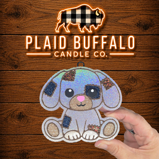 Hand-painted patchwork puppy freshie with iridescent white glitter, black and plaid fabric-style patches, tan glitter accents, a pink nose, and brown outlines, displayed on a rustic wood background.